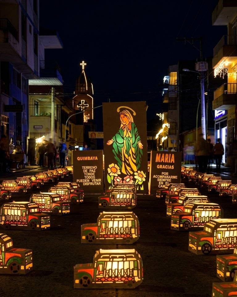 Cientos de faroles de papel iluminados en forma de chivas tradicionales y otros diseños llenan una calle nocturna en Quimbaya, Colombia. Al centro de la exhibición, una gran imagen iluminada de la Virgen María flanqueada por dos letreros artesanales con texto. Al final de la calle, se ve una iglesia iluminada.