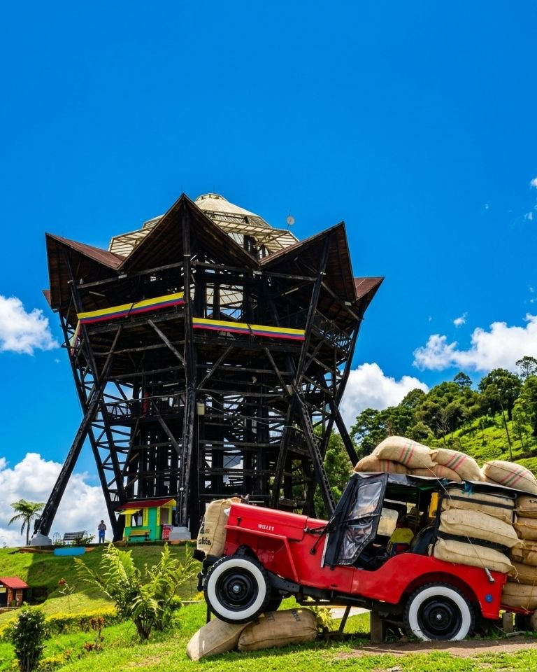 Un Jeep Willys rojo vintage, cargado en exceso con sacos de café de arpillera, estacionado en una ladera de hierba verde. Detrás, a la izquierda, se alza una gran y elaborada torre de observación de madera oscura con banderas de Colombia en sus niveles, bajo un cielo azul vibrante con nubes blancas dispersas.
